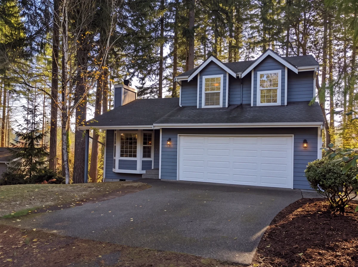 A two-story house with blue lap siding, twin gabled dormers and a two-car garage with the house number beside it, set among tall evergreens