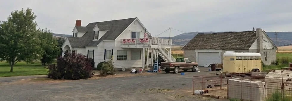 Earl's white farmhouse in Malin, Oregon, with gable dormers and a steep roof, beside a detached garage with a failing roof, photographed from the road in 2016