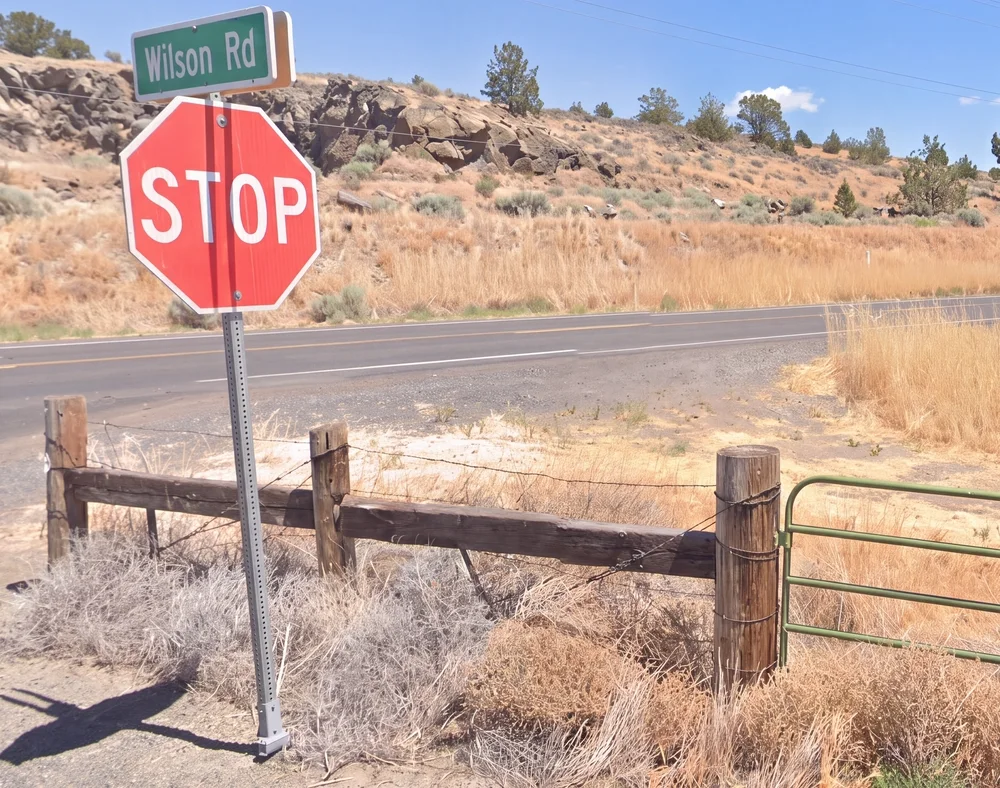 A green Wilson Rd street sign above a stop sign at a rural crossroads outside Malin, Oregon, with a wood-and-wire fence, dry grass and rimrock behind it