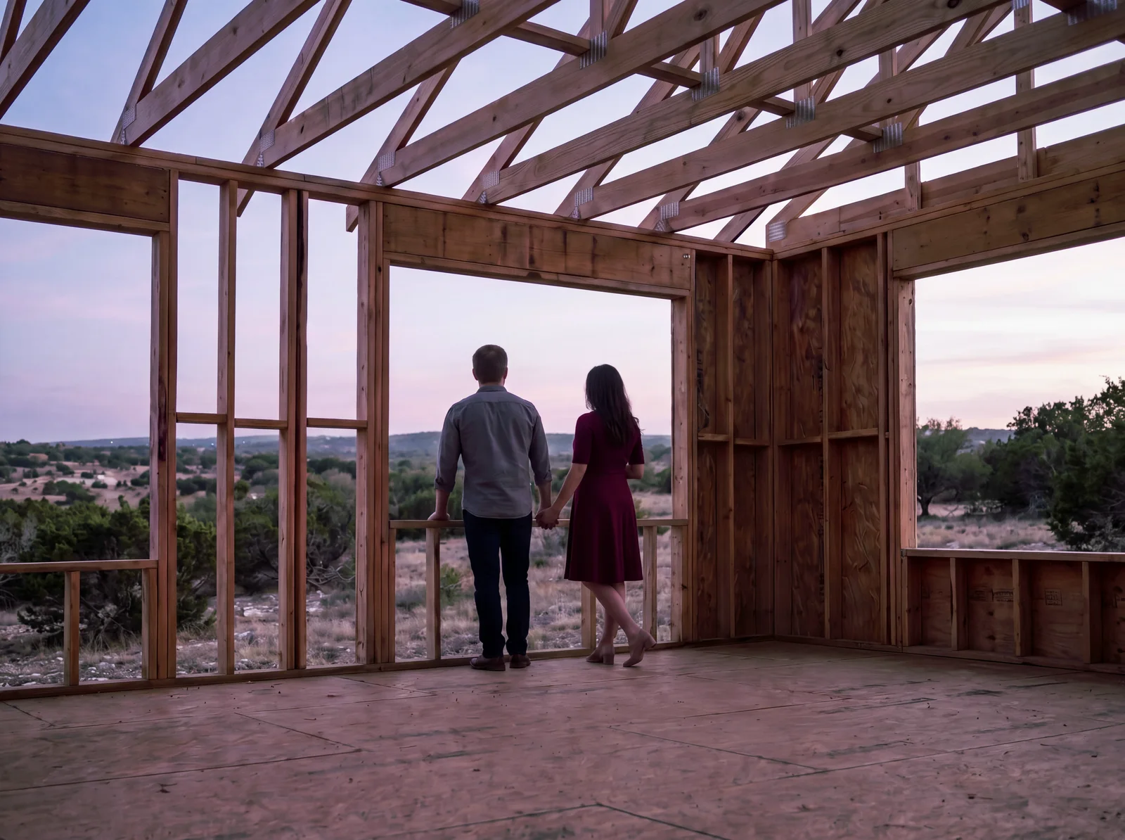 A couple standing hand in hand on the plywood subfloor of their half-framed house at dusk, looking out through an unfinished window opening at the Hill Country